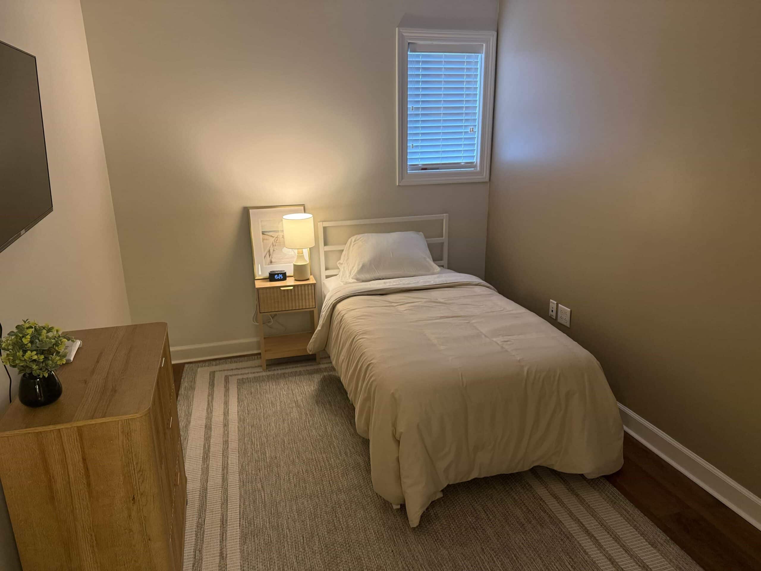A room with a made-up bed, a small window to the right, and a small table with a mirror to the left.