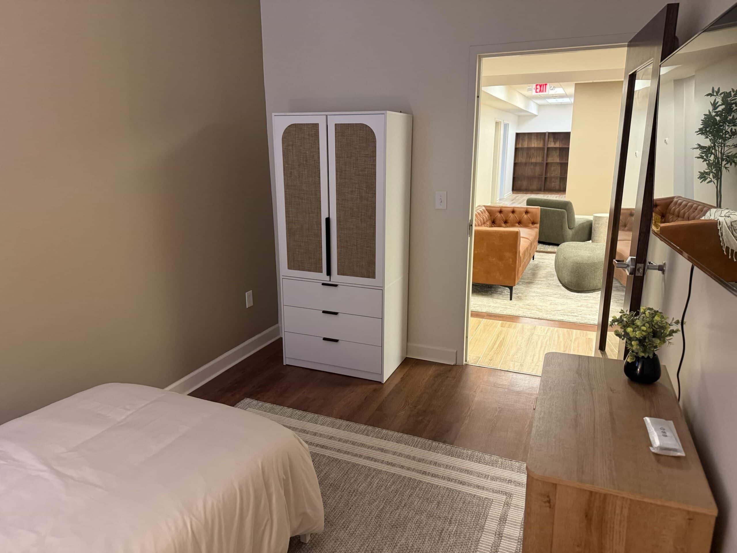 Part of a made bed, a small wardrobe, a table, and a mirrored door leading to the living room.